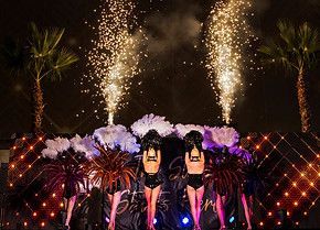 Nighttime stage show with dancers and fireworks. Palm trees frame the stage. Dancers in black costumes.