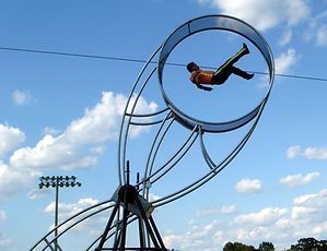 A person performing a stunt inside a large metal wheel against a blue sky.