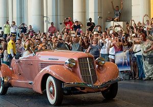 Pink vintage car driving, surrounded by a crowd of cheering people.