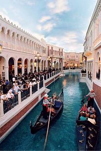 Canals with gondolas in a Venetian-style setting with people on balconies, blue water, and pale buildings.