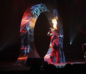 Fire breather in red outfit performs under an arch with projected images, stage lit.