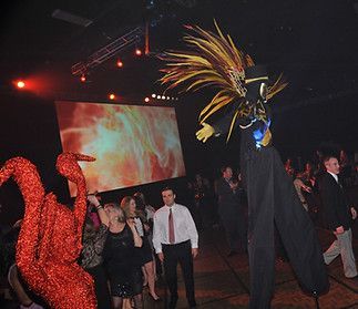 Party scene with a stilt walker in a feathered headdress and a red, abstract figure interacting with guests.