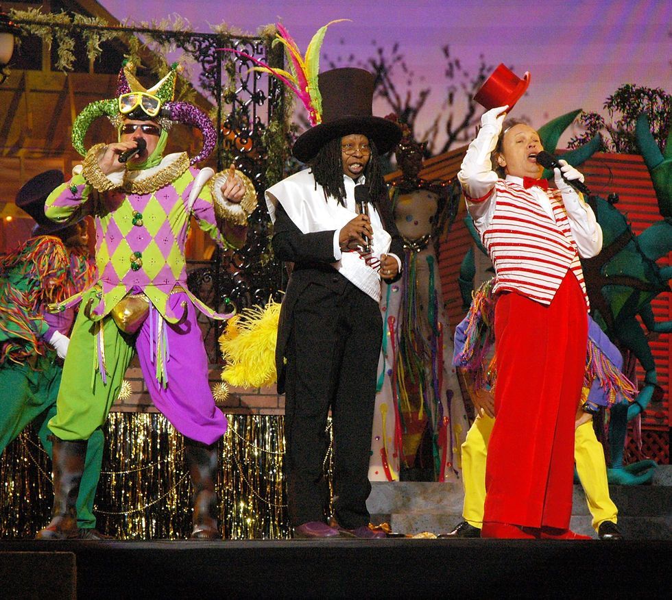 Men in colorful costumes, Mardi Gras style, on stage: performer with top hat, jester, and one singing.