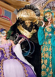 Three people in ornate masks and costumes, posing in a grand, gilded room.