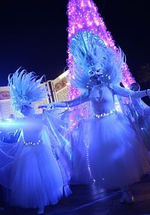 Two dancers in white costumes with feathery headdresses perform at night with a purple lit tree in the background.