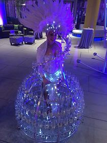 Woman in illuminated champagne glass skirt with feather headdress, serving drinks at an event.