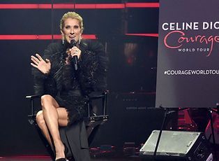 Celine Dion sits onstage, speaking into a microphone. She wears black and waves.