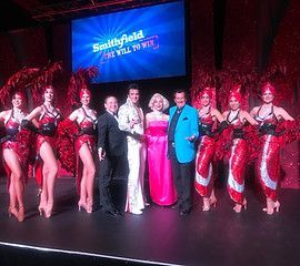 A group of performers posing onstage. Includes Elvis, Marilyn, showgirls, and men in suits under a