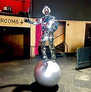Person in silver costume balances on a large silver ball, pointing to the left.