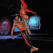 Two acrobats on a rope: man supporting woman, both in red and black outfits; stage with