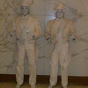 Two people in white suits and makeup stand still against a marbled wall.