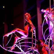 Performers in a cabaret show, using metal structures as props, lit with purple and orange lights.