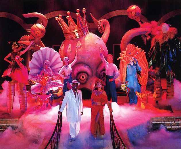 Stage with costumed performers, large octopus prop wearing a crown, bathed in pink light.