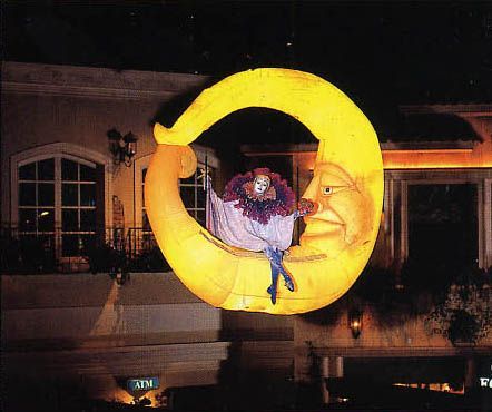 Large, illuminated crescent moon with smiling face, holding a clown. Outdoor setting, night.