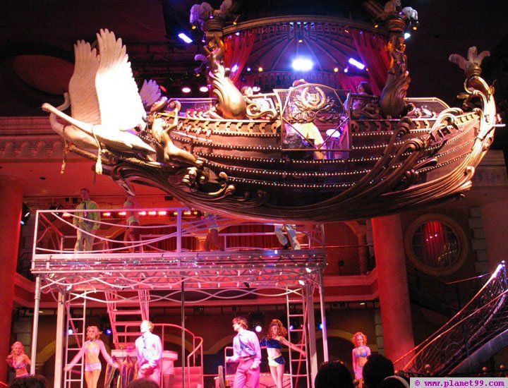 A stage production with performers under an ornate ship prop, bathed in red and blue stage lights.