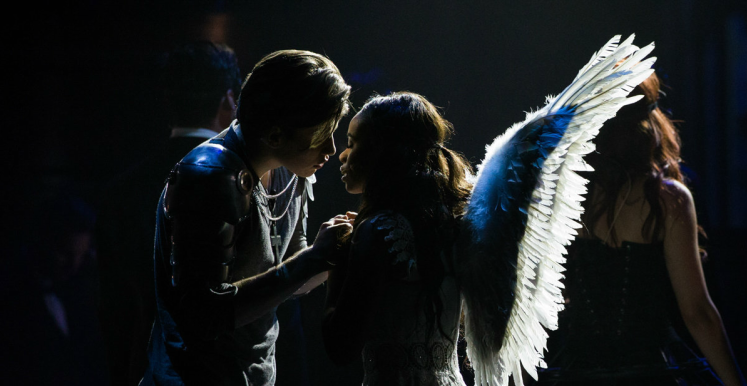 Man in silver jacket leans toward woman with angel wings in spotlight.