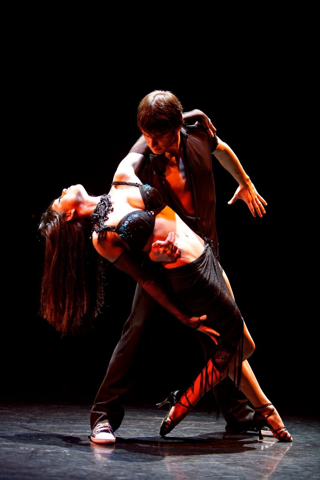 Man and woman in passionate dance pose on dark stage. He supports her as she arches back in black dress.