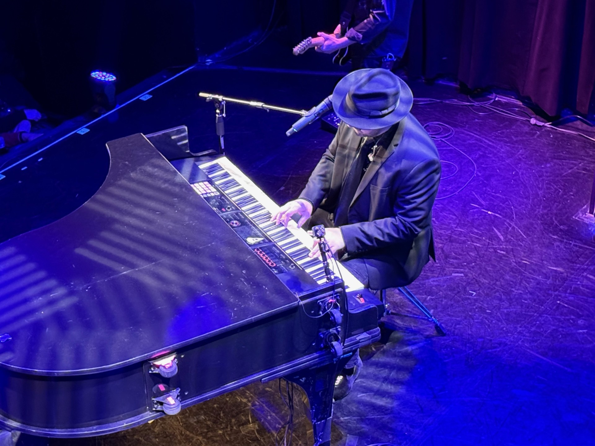 Man in a fedora playing a grand piano on stage, bathed in blue light.