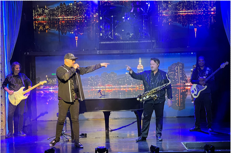 A band performs on stage with a New York City backdrop. A singer points, saxophonist plays, others with guitar and bass.