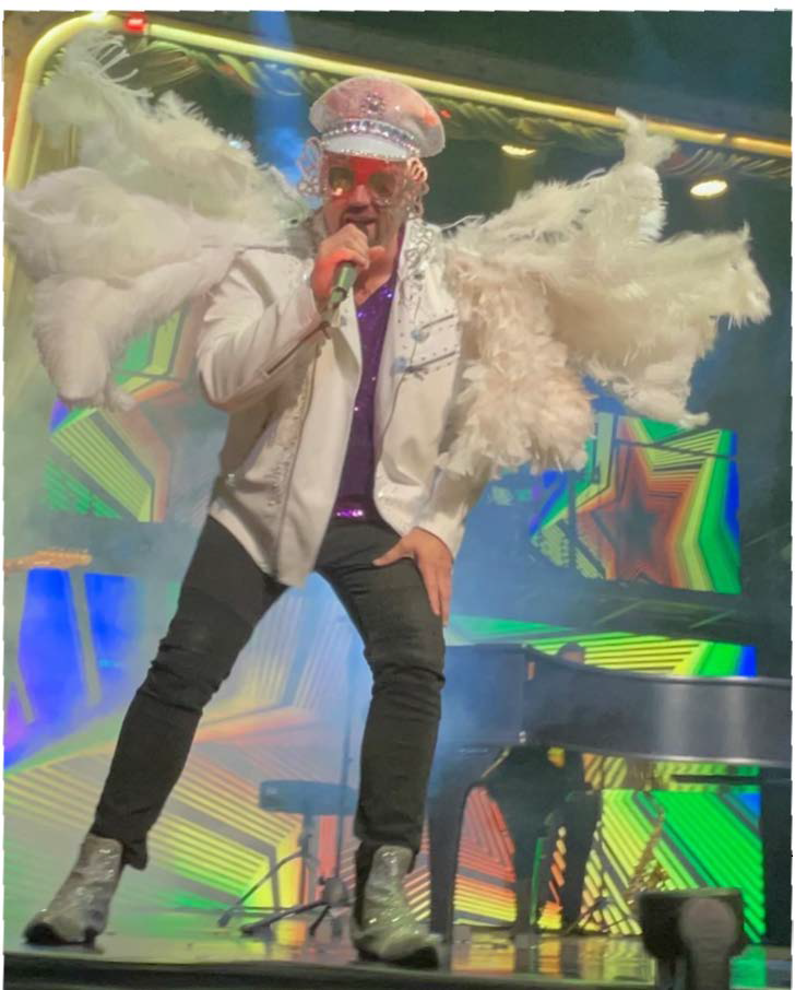 Man in white jacket and feathered wings singing on stage, next to a piano.