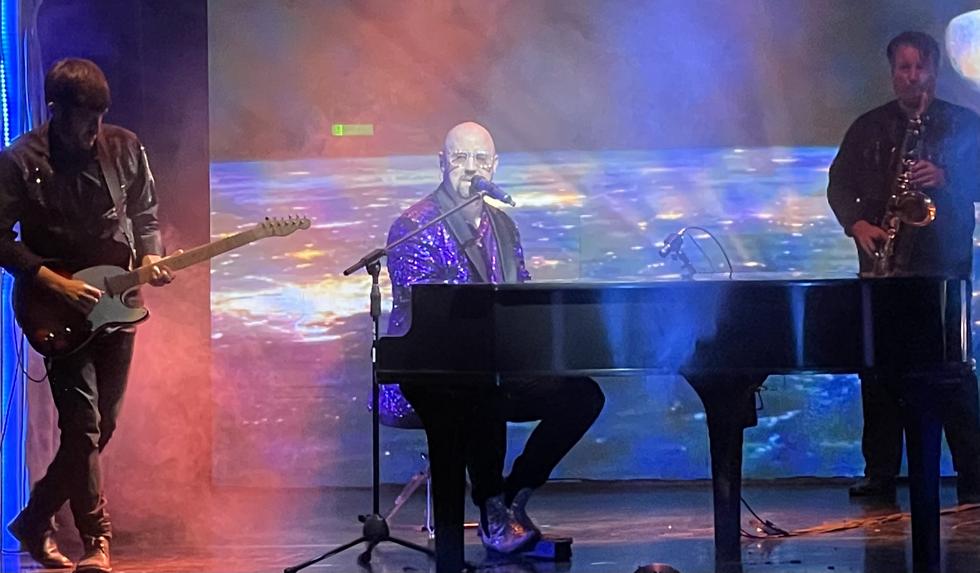 A bald musician in a purple suit plays piano and sings, flanked by a guitarist and saxophonist on stage.