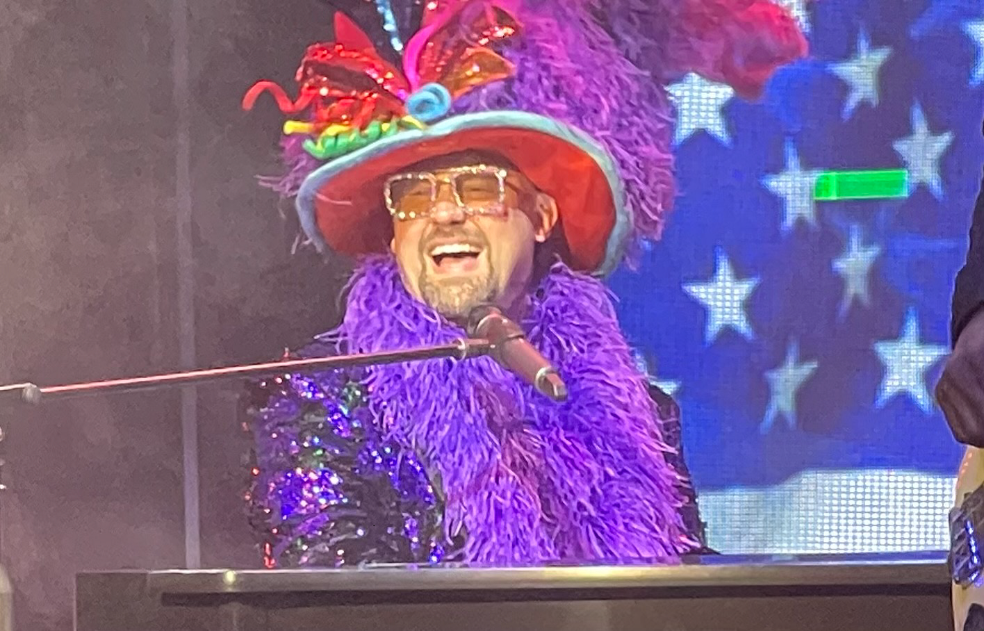 A man, Elton John impersonator, in purple feathered outfit, laughing at piano, stage background with American flag.