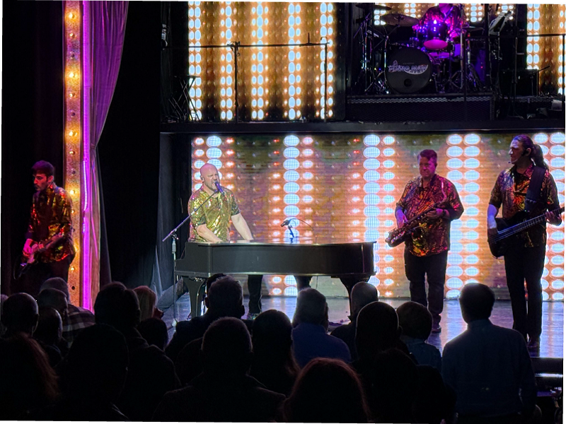 A band performs onstage: piano player, saxophone, guitar, drums, colorful lights, and audience.