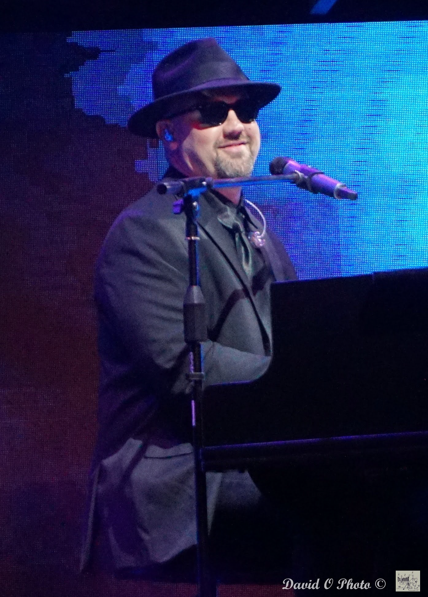 Man in hat and sunglasses at a piano, singing into a microphone on a stage.