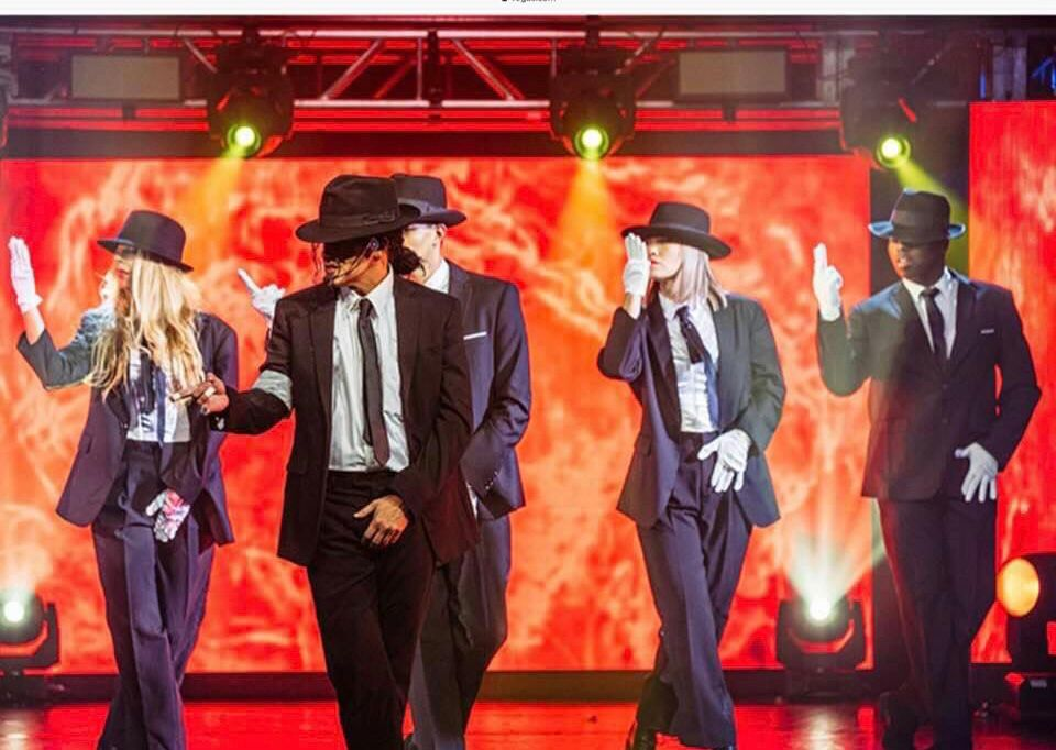 A group of performers dressed in black suits, fedora hats, and white gloves dance on a red stage.