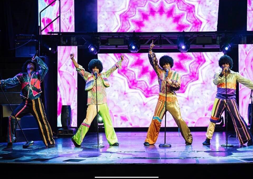 Four performers in colorful 70s outfits sing on stage with a psychedelic backdrop.