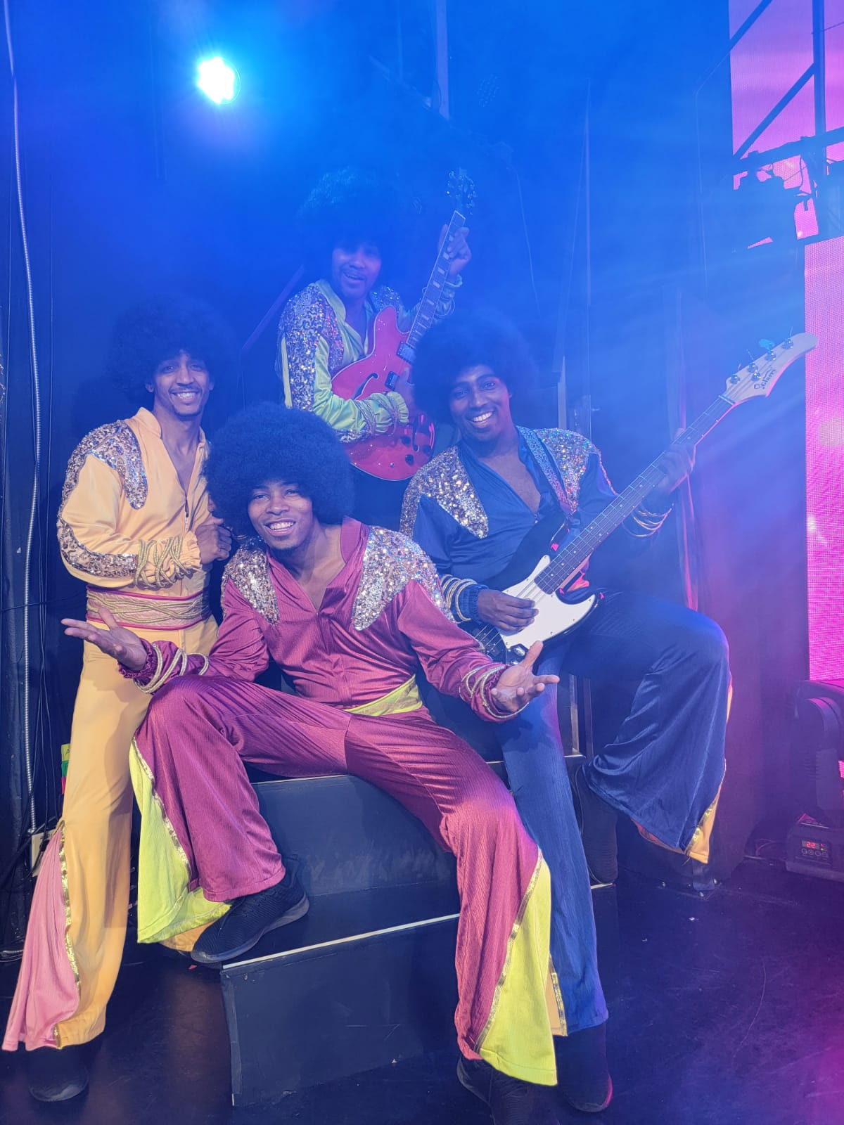 Four people in colorful 70s attire, playing instruments. Purple stage with blue lighting.