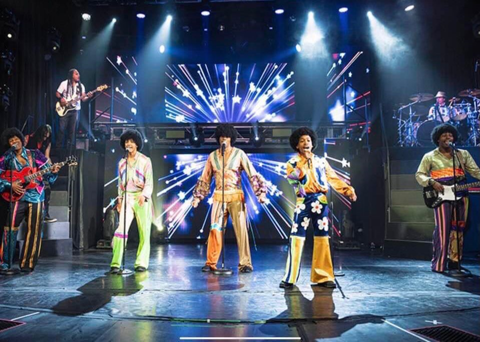 Band on stage, performing. Five vocalists in colorful outfits, musicians, and a backdrop with lights.