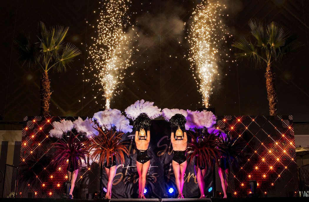 Stage show with dancers, fireworks, palm trees, and decorative lights.