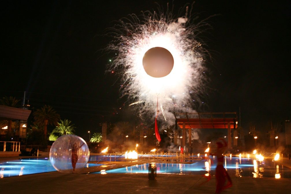 Nighttime fireworks display over a pool, resembling an eclipse. Performers with fire torches and in a bubble.