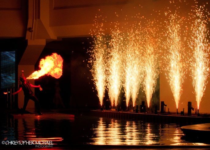 Fire breather on stage, with orange flames, water, and fireworks.