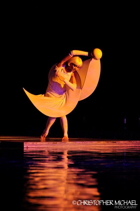 A performer in a crescent moon costume on a reflective stage; yellow, purple, and brown.