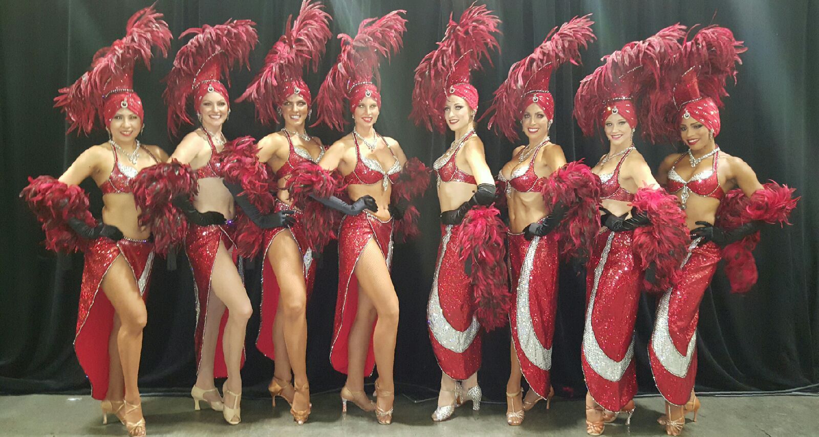 Nine showgirls in red costumes with feathered headdresses pose for the camera.