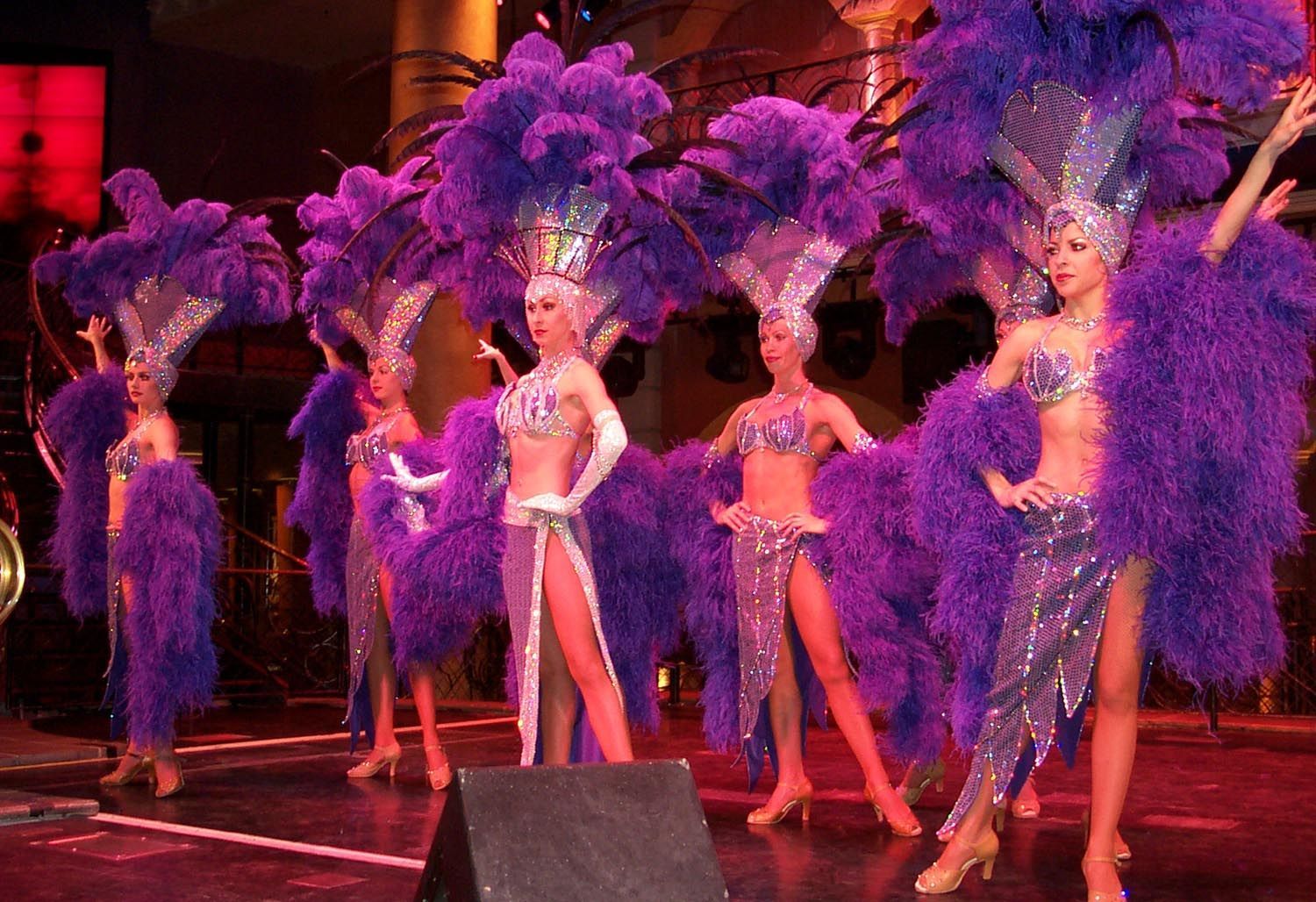 Showgirls in purple feathered headdresses and costumes perform on stage.