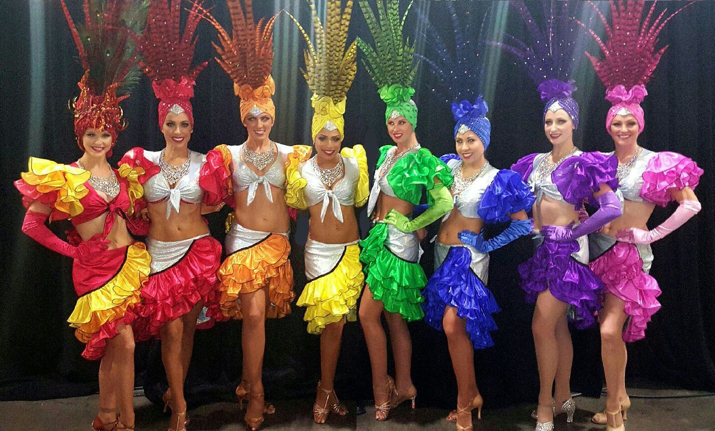 Seven showgirls in rainbow-colored costumes, each with elaborate feather headdresses, posing in a dark setting.