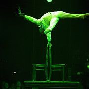 Acrobat in a green-lit performance, balancing on one hand atop stacked chairs, legs split in a