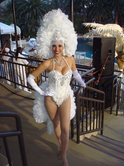 Woman in a white showgirl costume smiles, near a pool; Vegas setting.