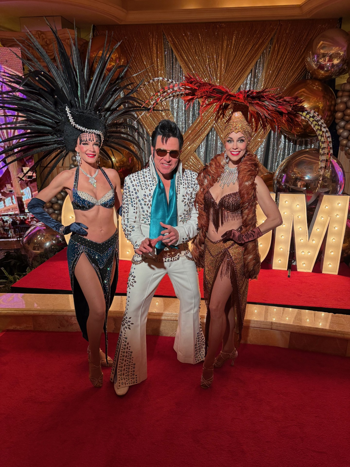 Elvis Presley impersonator with two showgirls on a red stage with a 