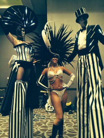 Three costumed performers, two on stilts. Black and white striped suits. One woman in a bikini stands in the center.