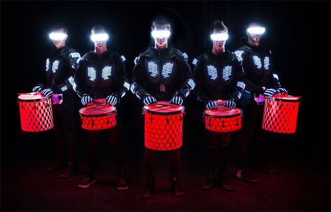 Drummers in black illuminated outfits and glowing red drums. Dark stage with neon lights.