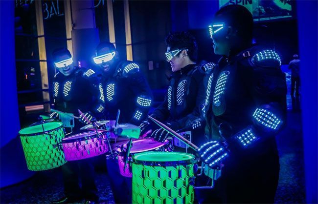 Drummers in black suits with glowing blue lights perform at night, drums in neon green and pink.