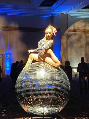 A dancer in a sparkly bodysuit on a large disco ball, performing in a ballroom.