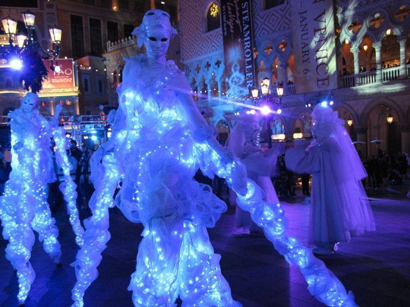 Performers in glowing blue costumes at an outdoor nighttime event in front of ornate buildings.