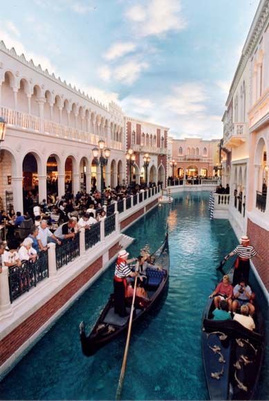 Gondolas on a canal in a Venetian-themed shopping area, people dining at outdoor restaurants.