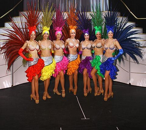 Seven showgirls in colorful feather headdresses and costumes, posing on a stage.