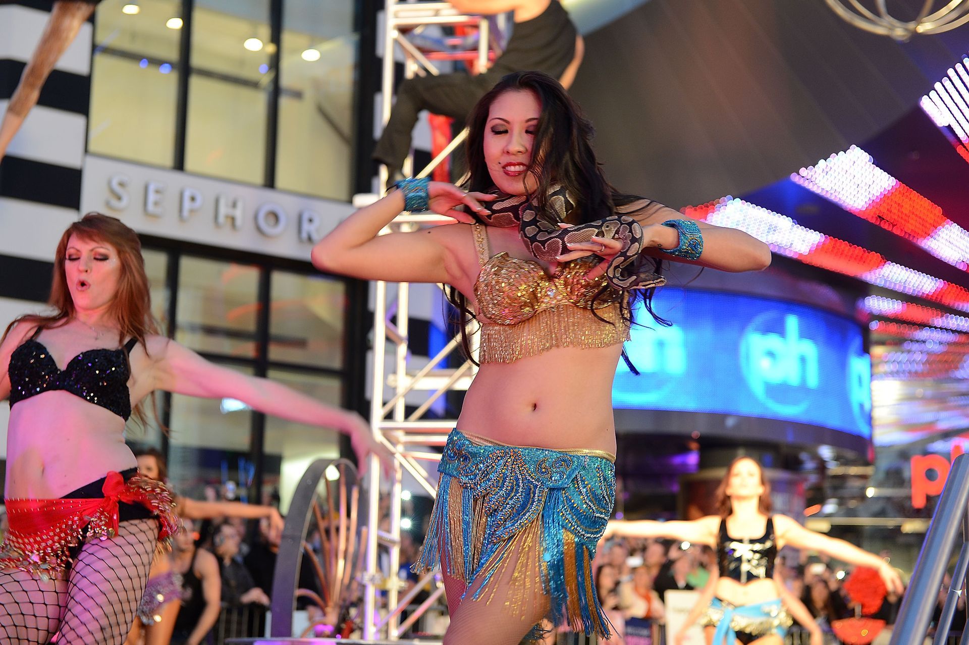 Women belly dancing on an outdoor stage at night, in front of a large screen and a Sephora store.